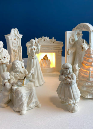 White Porcelain Figurines of Santa & His Family Celebrating Christmas in Front of Fireplace. Illuminated Fireplace and Tree. Mantel Display.